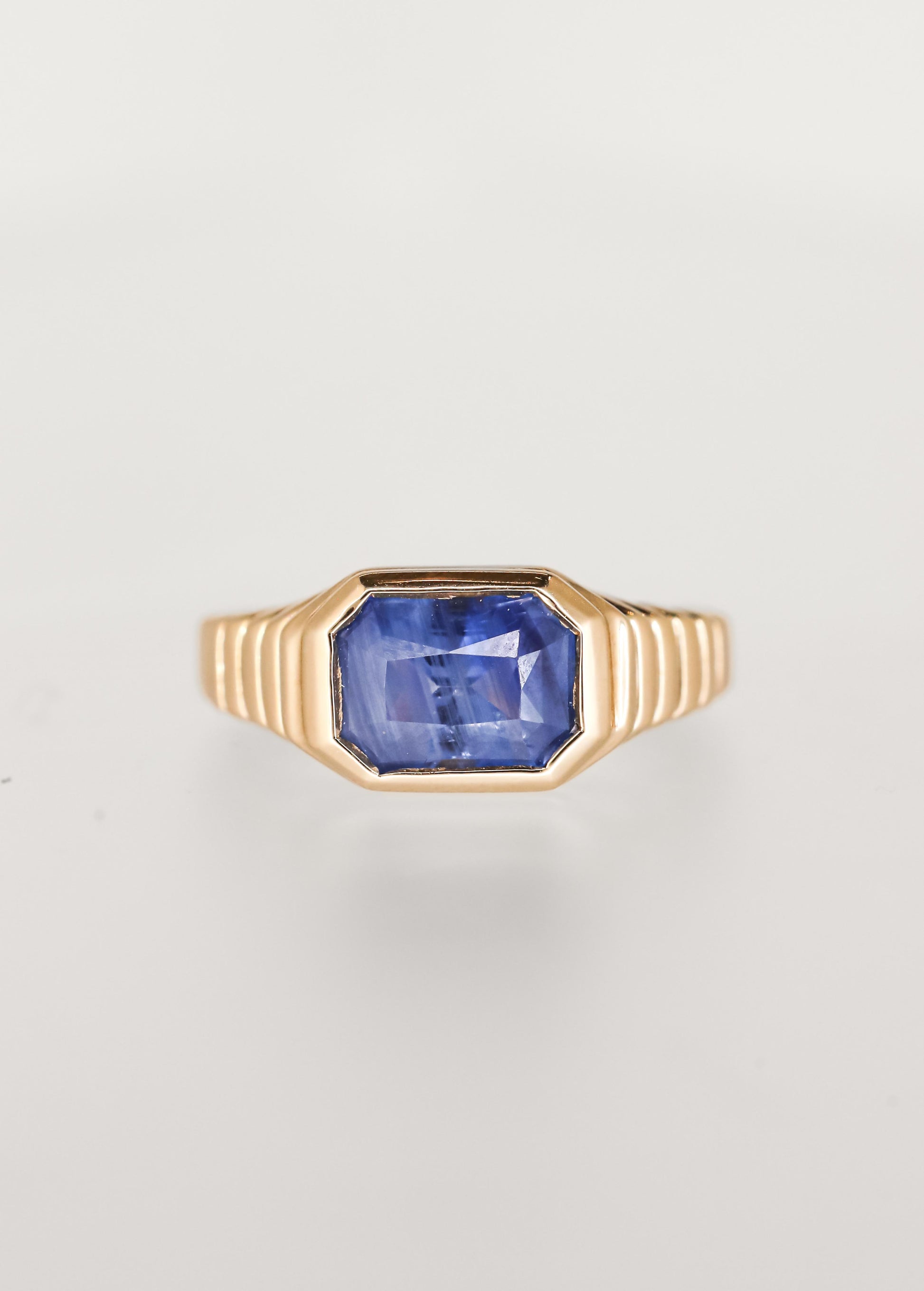 Masculine leaning east west emerald cut blue sapphire engagement ring in 14K yellow gold with a sculptural ribbed signet bezel