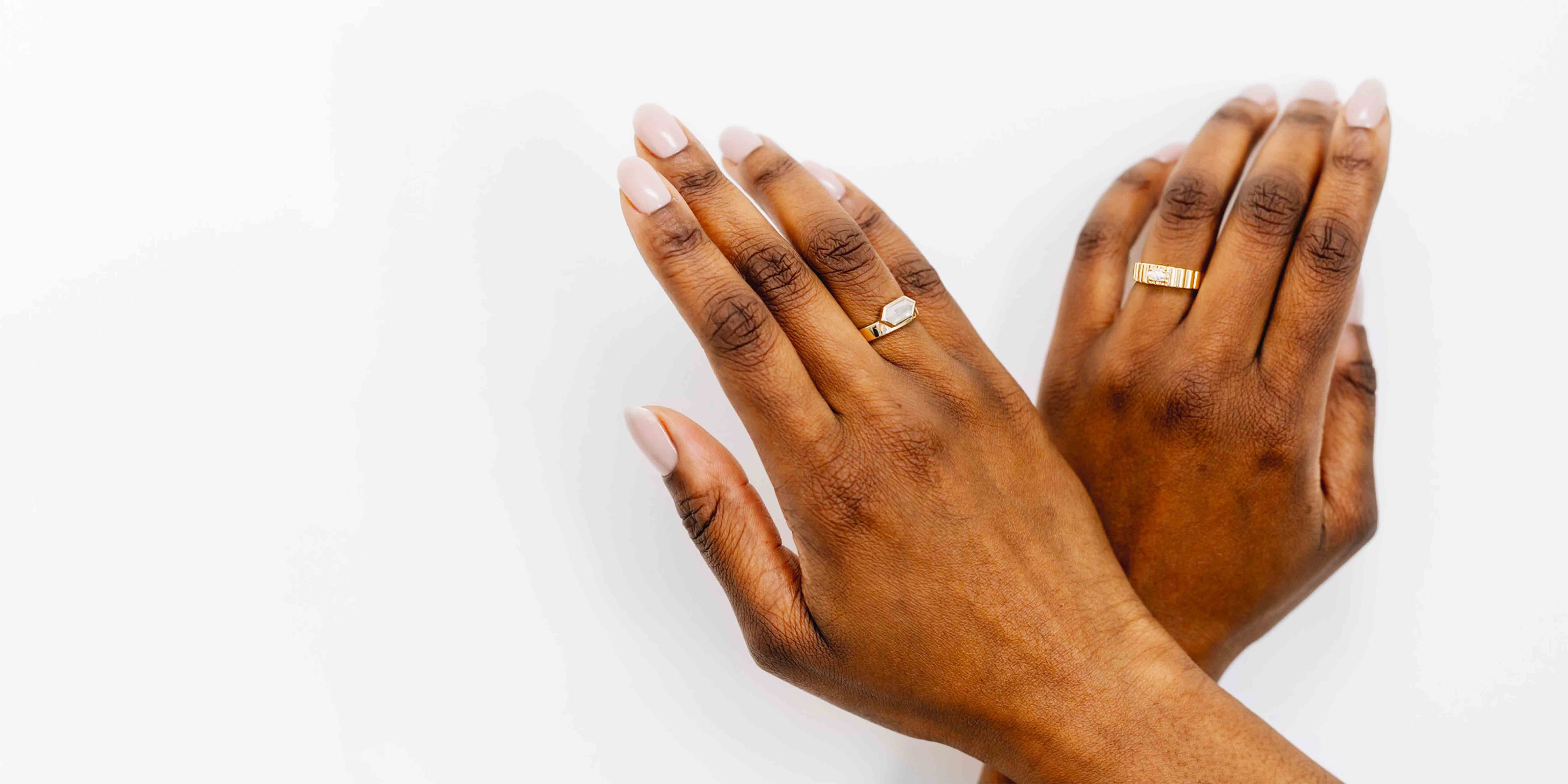 Image of two hands resting on a white surface, each adorned with a ring from a gender-neutral collection. One hand wears a minimalist hexagonal-cut salt and pepper diamond bezel set in a yellow gold band, while the other showcases a ribbed gold band featuring a small east-west set oval moissanite. Both hands have neatly manicured, light pink nails, emphasizing the subtle elegance of the jewelry.