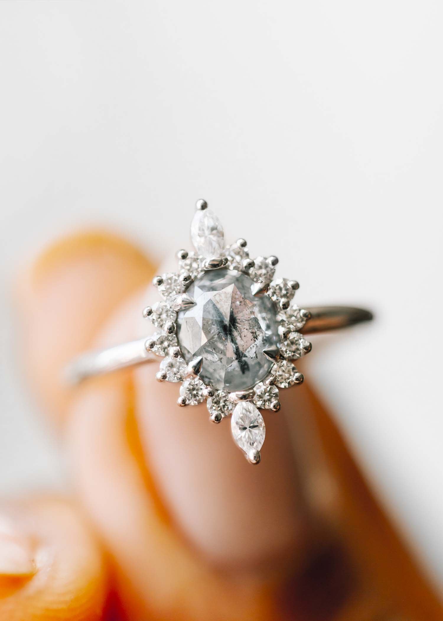 A close-up image of a unique engagement ring featuring an oval salt and pepper diamond at its center. The diamond is surrounded by a halo of sparkling white diamonds and accented with marquise-cut diamonds, creating a floral-inspired design. Set in a white gold band, this artisanal ring showcases the striking contrast and natural beauty of the salt and pepper diamond. Perfect for those seeking distinctive, non-traditional engagement rings.