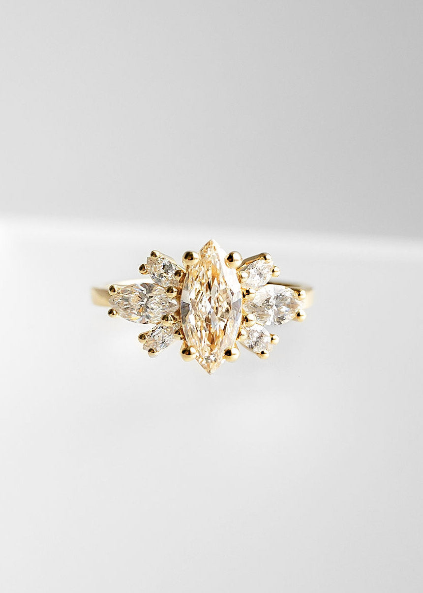 Exquisite gold engagement ring featuring a central marquise-cut champagne diamond surrounded by various sizes of marquise-cut clear diamonds, set in a radiant sunburst pattern. This design blends vintage charm with modern elegance, showcased against a soft grey background.