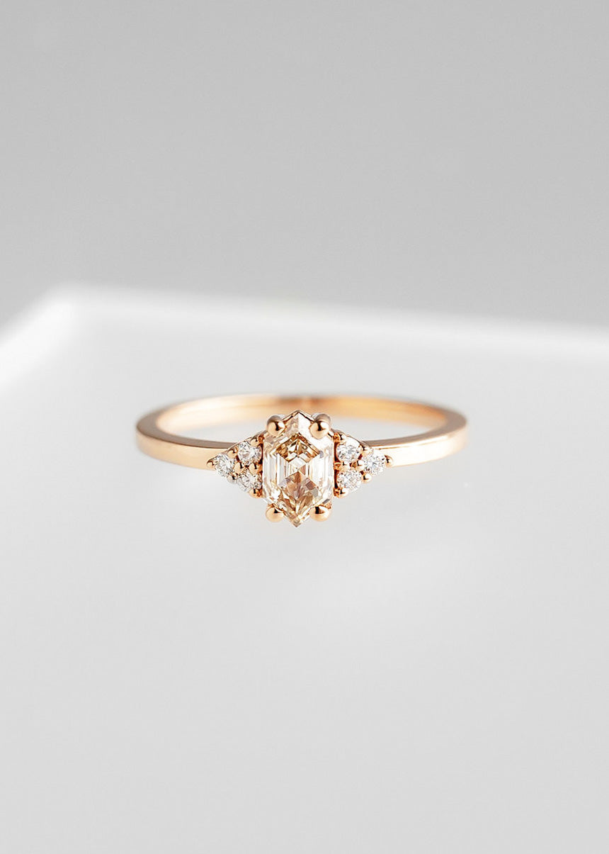 Delicate rose gold engagement ring featuring a central hexagon-cut champagne diamond, elegantly accented by three round-cut diamonds arranged in a triangular shape on each side. The design creates a harmonious symmetry and balance, showcased against a soft grey background to emphasize the refined and sophisticated appeal of the ring.