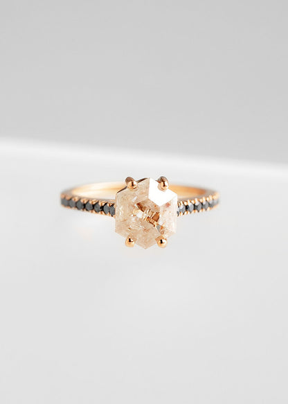 Elegant rose gold engagement ring featuring a prominent rose-colored salt and pepper diamond in a unique hexagonal cut, prong set to enhance its natural beauty. The band is adorned with a row of delicate black diamonds, adding a modern contrast to the overall design, all showcased against a soft grey background.