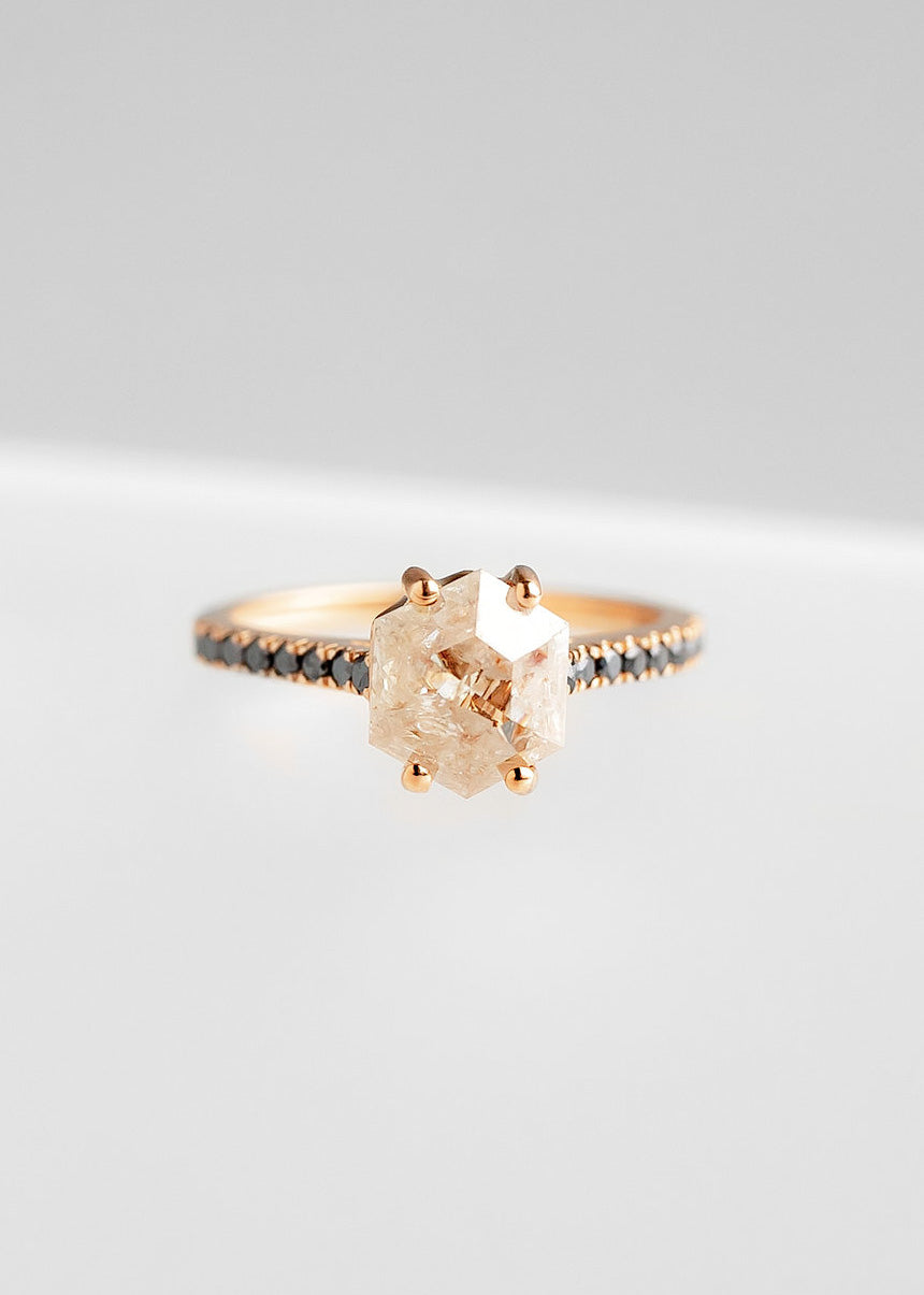 Elegant rose gold engagement ring featuring a prominent rose-colored salt and pepper diamond in a unique hexagonal cut, prong set to enhance its natural beauty. The band is adorned with a row of delicate black diamonds, adding a modern contrast to the overall design, all showcased against a soft grey background.