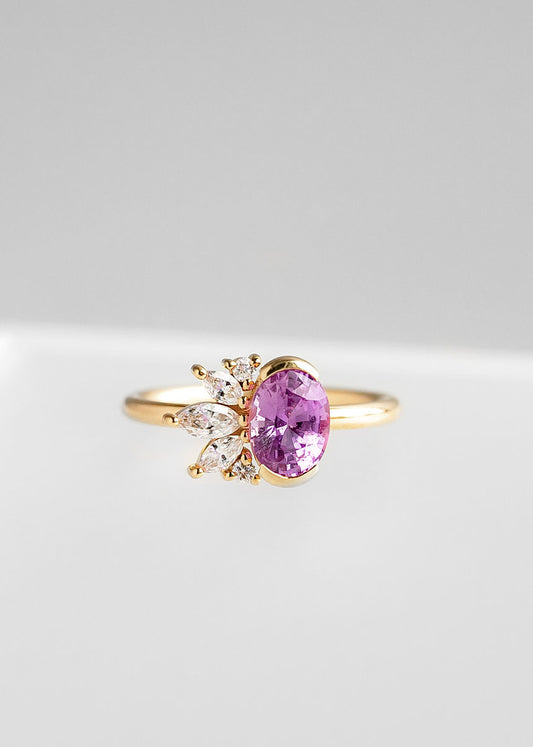 Elegant yellow gold engagement ring showcasing a vibrant oval-cut pink sapphire, beautifully complemented by a dynamic floral arrangement of marquise and round-cut diamonds. This design blends contemporary flair with traditional elegance, presented against a soft grey background to accentuate the ring’s striking color contrast and intricate detailing.