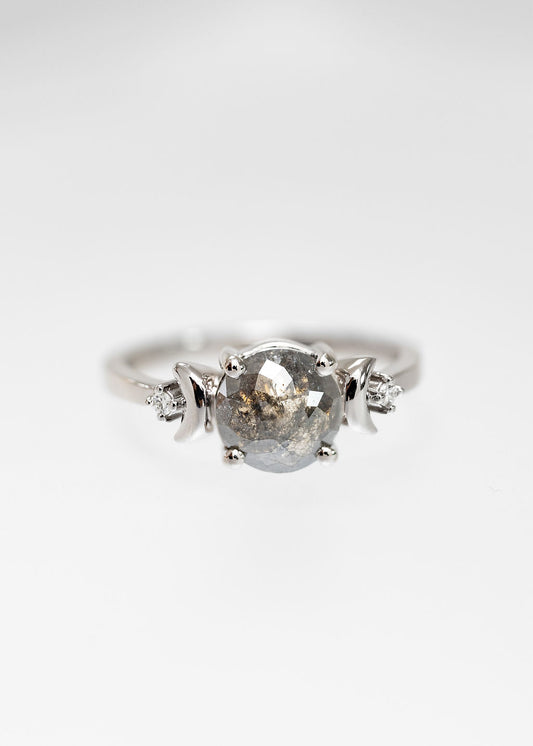 A close-up image of a unique engagement ring featuring a round salt and pepper diamond as the centerpiece. The diamond is set in a sleek white gold band and flanked by small, crescent moon-shaped accents and tiny round diamonds on either side. This artisanal ring combines modern elegance with celestial charm, making it an ideal choice for those seeking a distinctive and eye-catching piece. Perfect for anyone looking for a non-traditional, eco-friendly, and ethically sourced engagement ring.