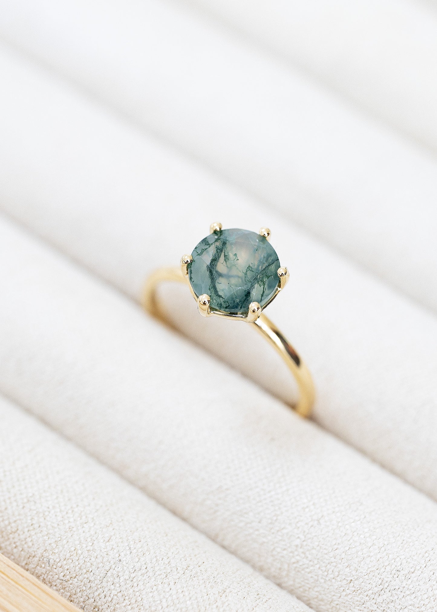 Nature-inspired engagement ring featuring a round moss agate center stone, securely held by six prongs in a classic yellow gold setting. The moss agate ring showcases unique green veining that evokes the look of forest moss, making it a standout choice for those who appreciate artisanal and organic jewelry designs. Presented on a textured fabric, this ring perfectly combines elegance with a touch of the natural world.